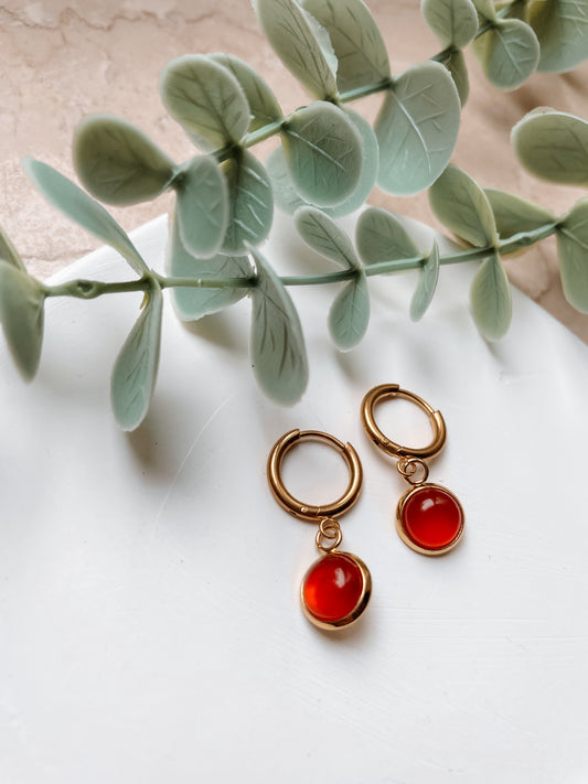Goldene Creolen-Ohrringe mit runden, roten Karneol-Steinen auf weißem Tablett mit Pflanzen