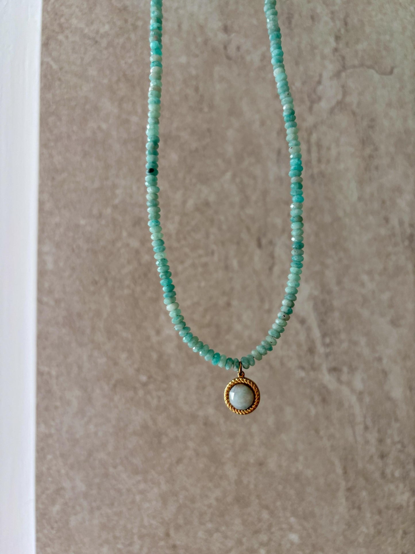 Blaue Amazonit-Perlenkette mit einem runden facettierten Amazonit Anhänger in Gold hängend vor grauer Wand