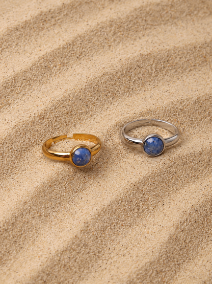 Zwei Lapislazuli-Ringe namens Amaya, einer in Gold und einer in Silber, liegen auf Sand.