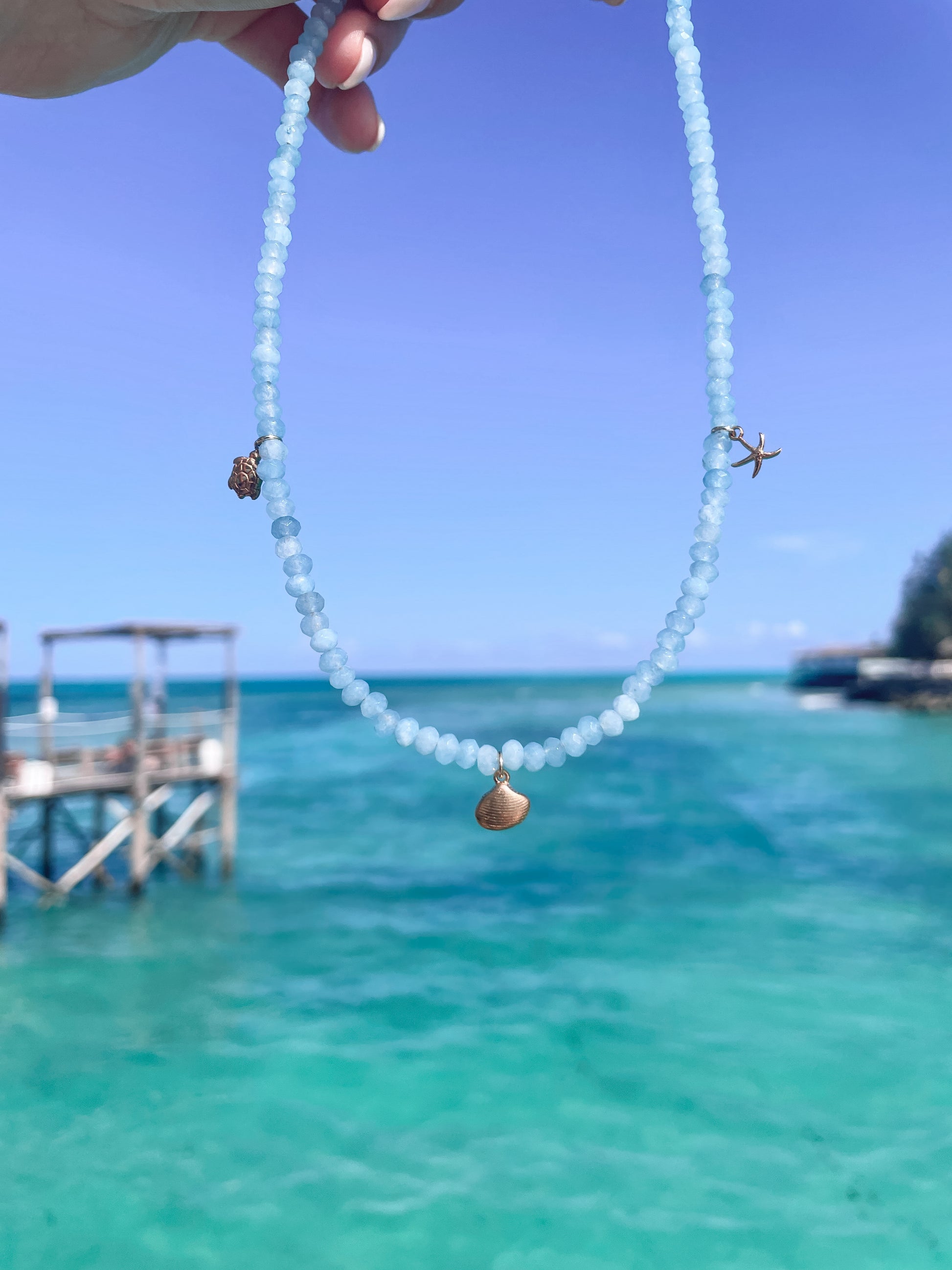 Blaue Jade-Perlenkette mit maritimen goldenen Anhängern hängend vor Meer