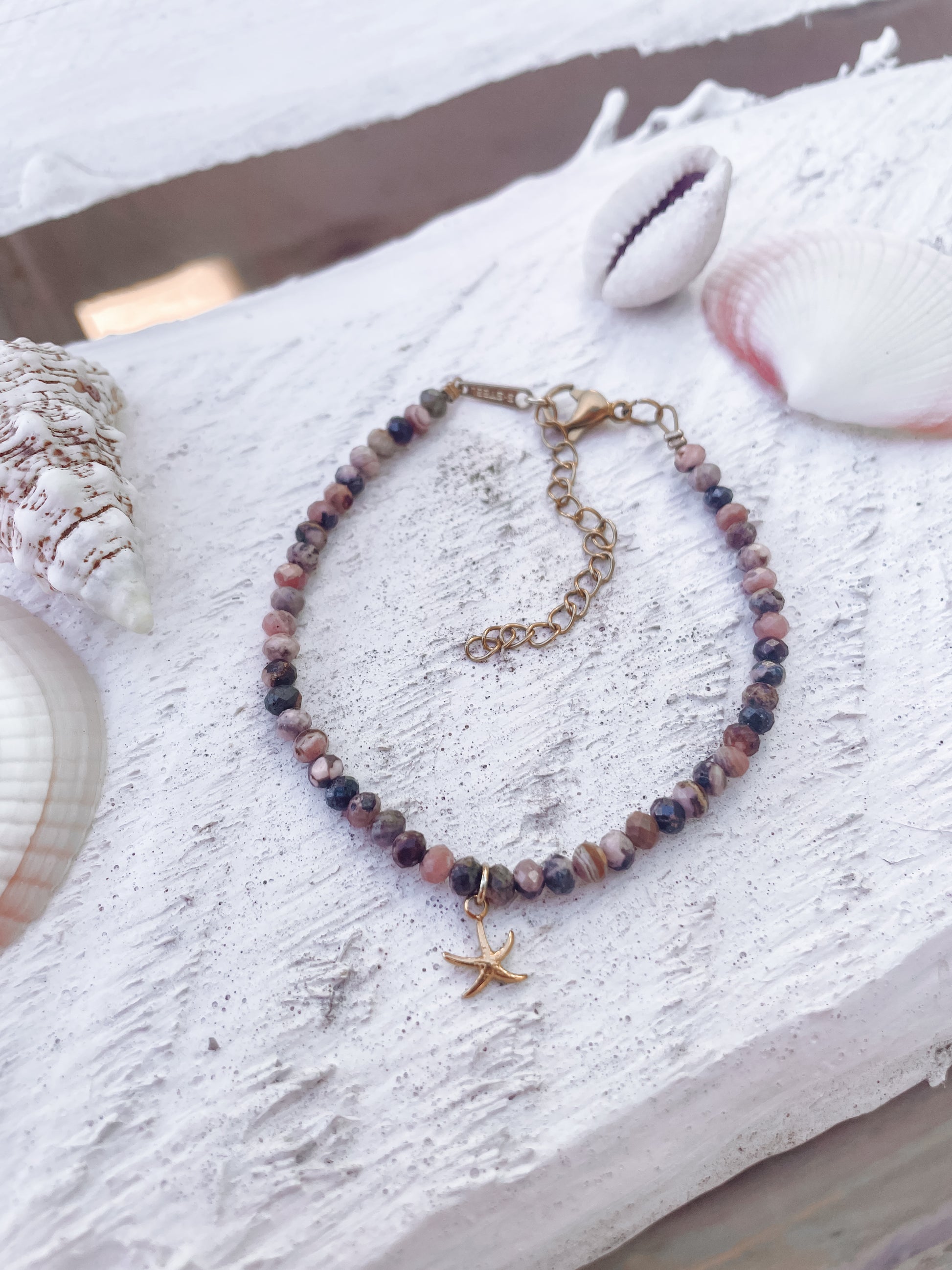 Rosa Braunes Rhodonit-Perlenarmband in Gold mit Seestern Anhänger liegend auf weißem Holz mit muscheln