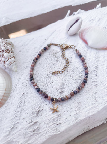 Rosa Braunes Rhodonit-Perlenarmband in Gold mit Seestern Anhänger liegend auf weißem Holz mit muscheln