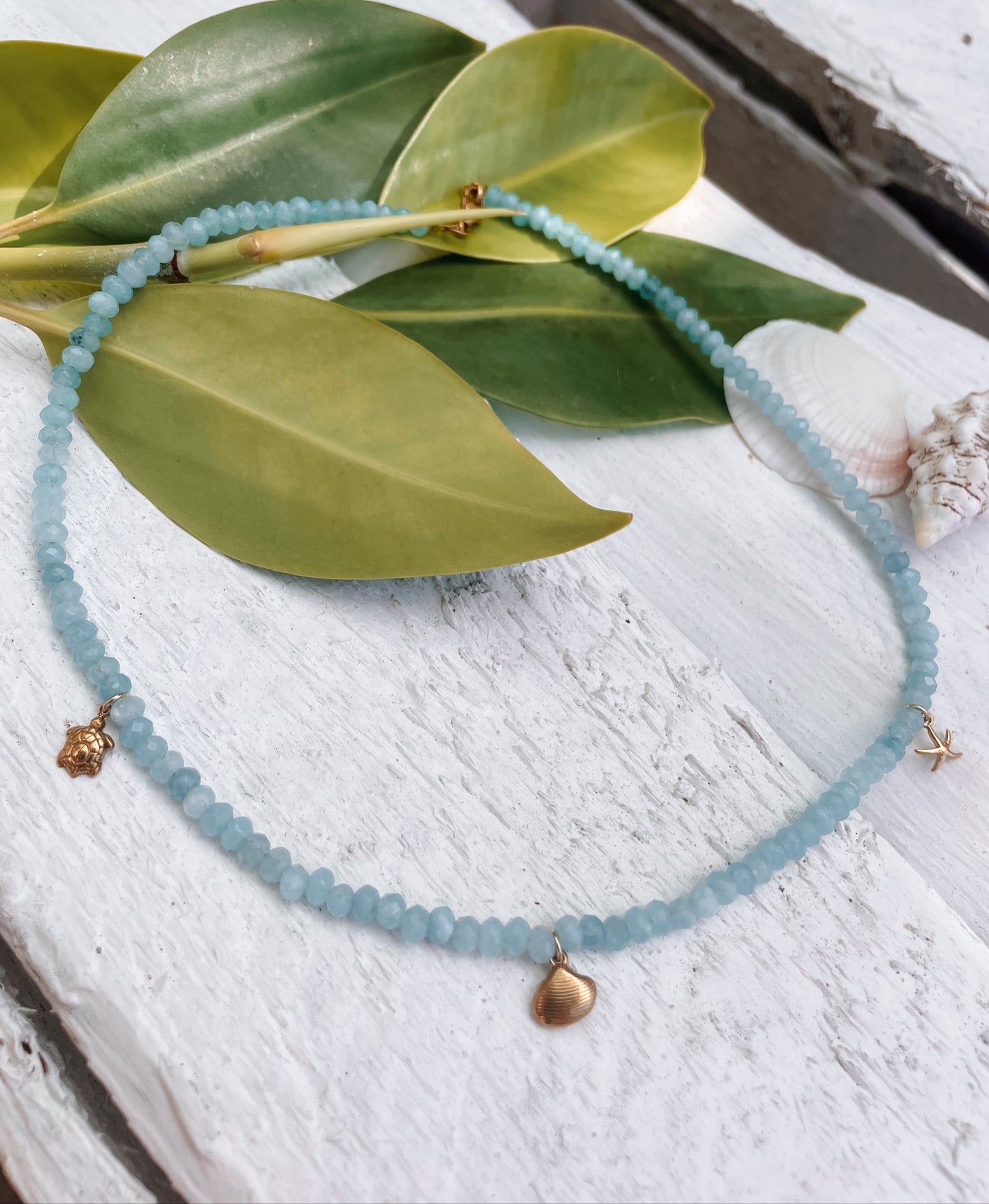 Blaue Jade-Perlenkette mit maritimen Goldanhängern liegend auf weißem Holz mit Pflanzen