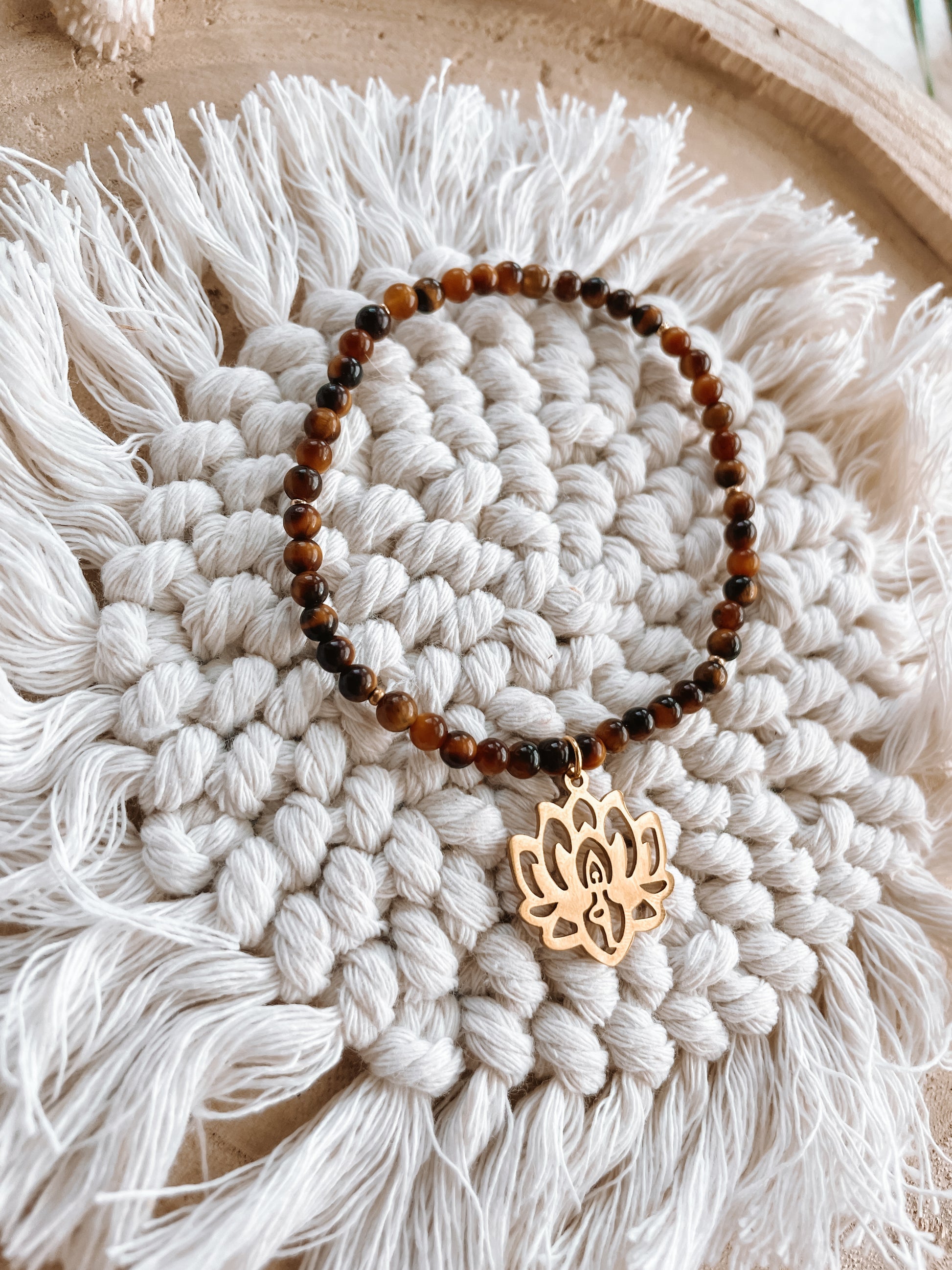 braunes Tigerauge-Perlenarmband in Gold mit Yoga Lotusblume Anhänger liegend auf Makramee Untersetzer
