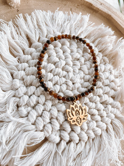 braunes Tigerauge-Perlenarmband in Gold mit Yoga Lotusblume Anhänger liegend auf Makramee Untersetzer