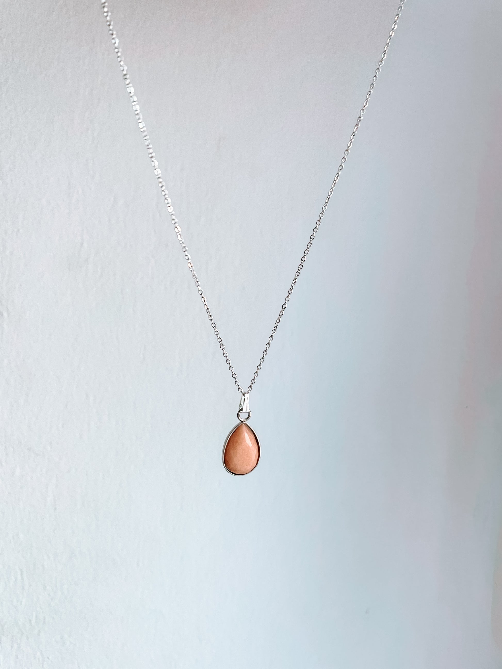 Handgemachte rosa Sonnenstein-Kette mit Tropfen Anhänger in Silber, hängend vor weißer Wand