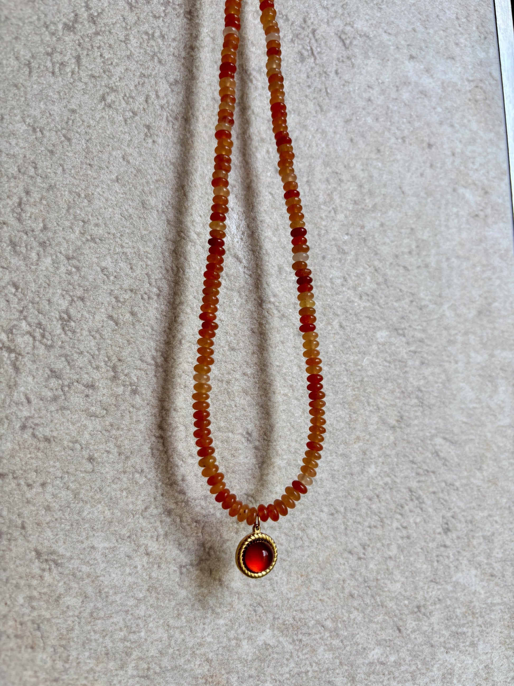 Handgemachte rote Karneol-Perlen-Kette mit rundem Anhänger in Gold, hängend an grauer Wand