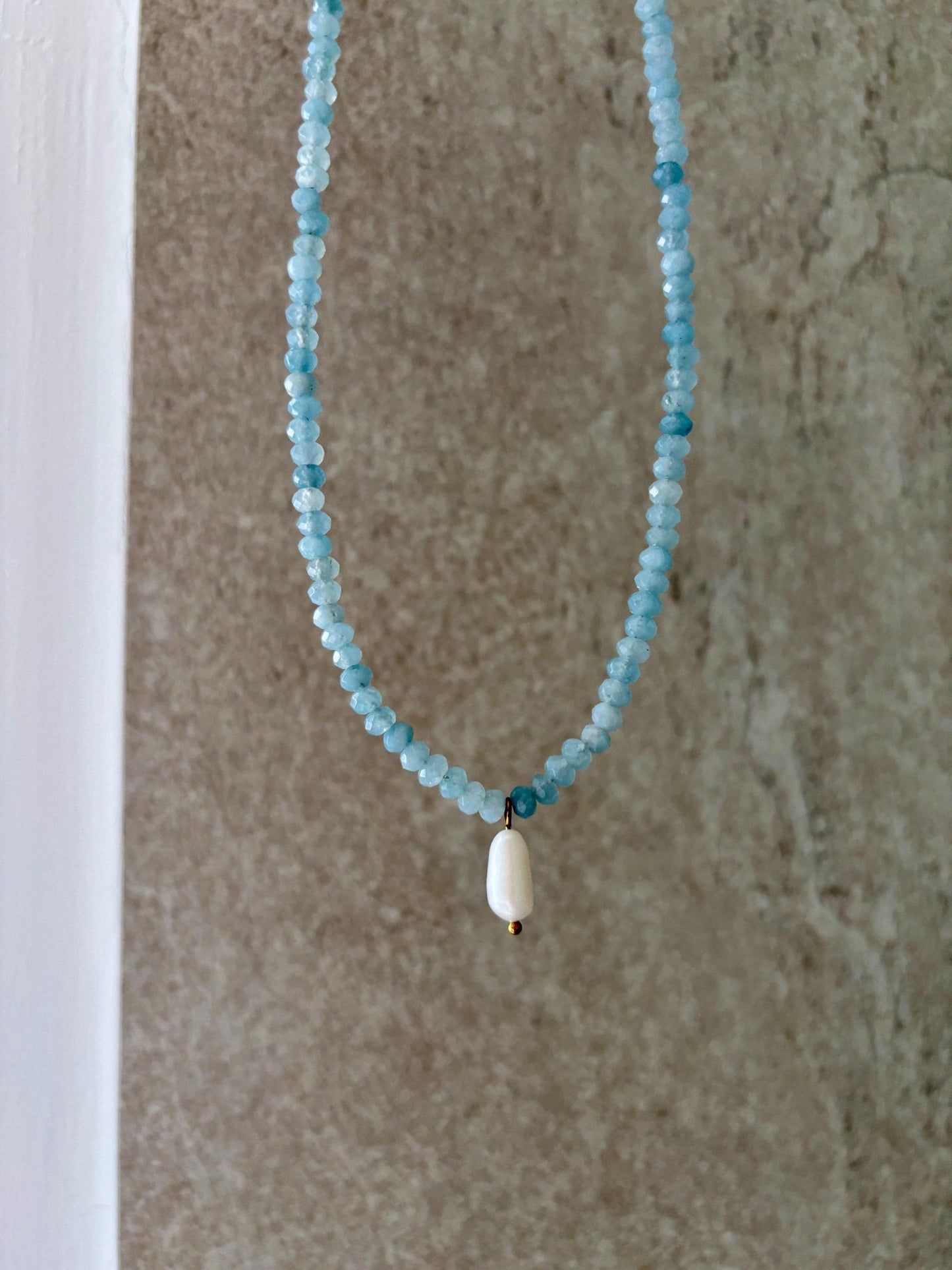 Blaue Jade-Perlenkette mit einem weißen Süßwasserperlen Anhänger hängend vor grauer Wand