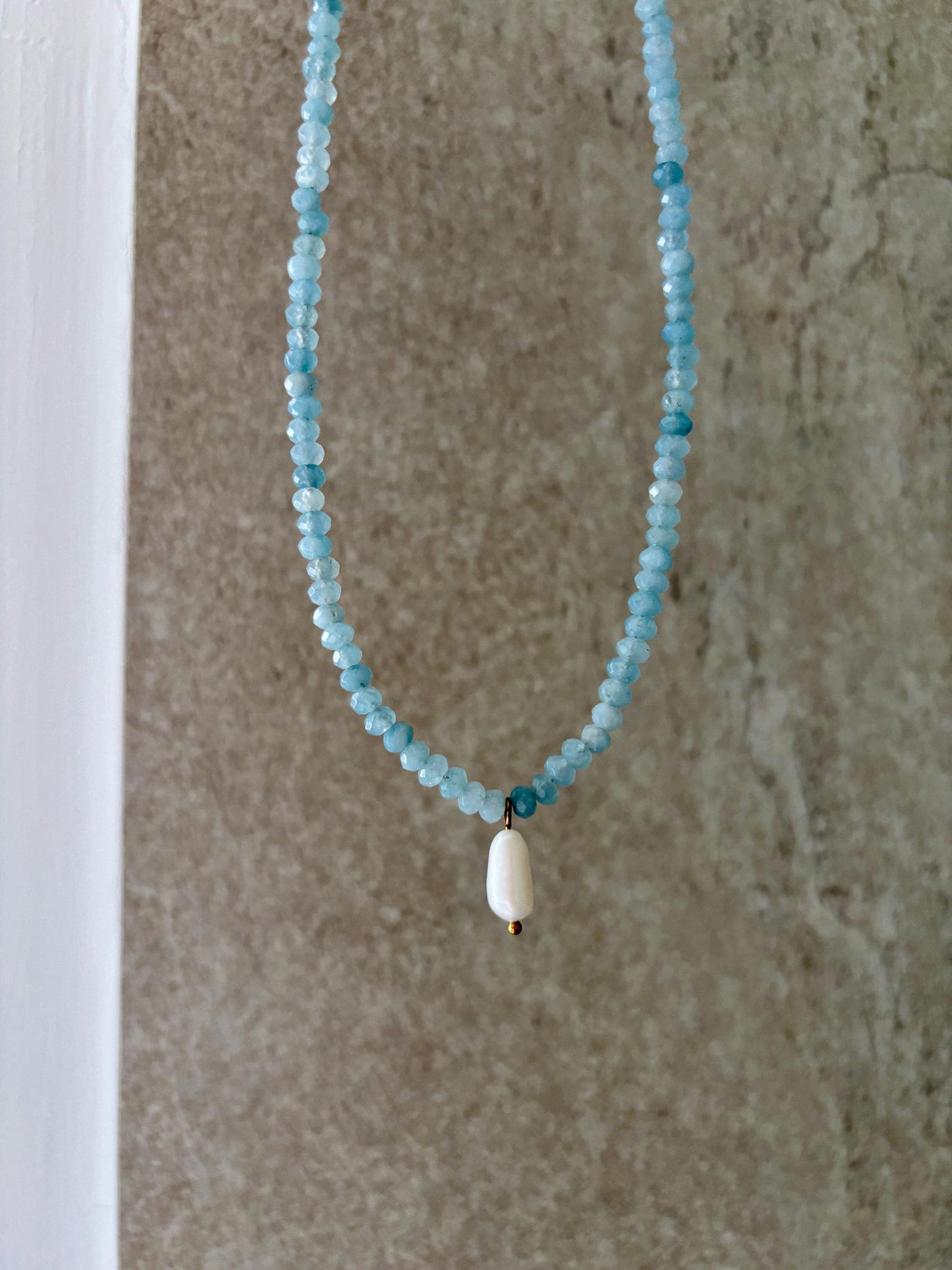 Blaue Jade-Perlenkette mit einem weißen Süßwasserperlen Anhänger hängend vor grauer Wand