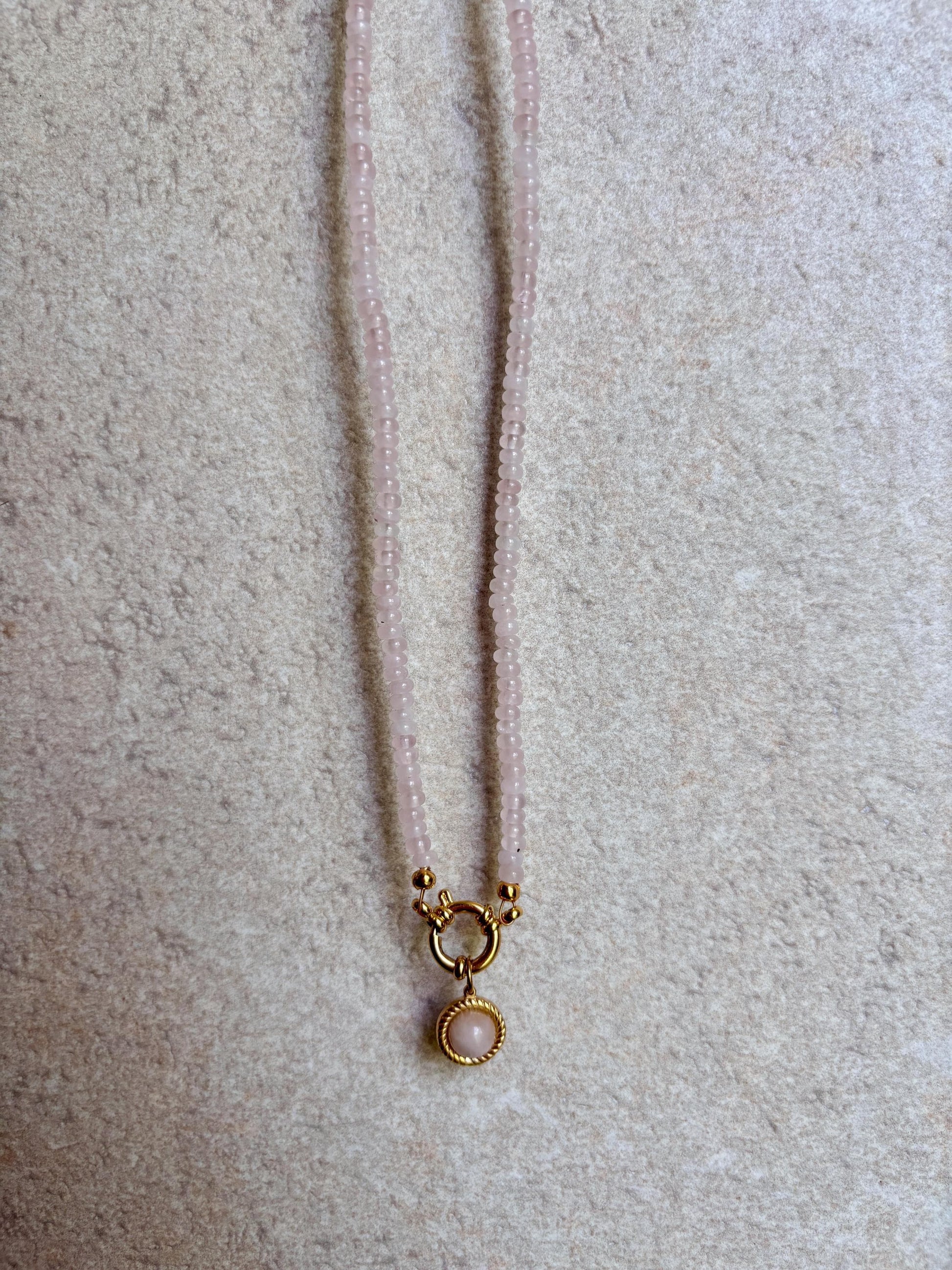 Rosenquarz-Perlenkette mit einem runden goldenen Rosenquarz Anhänger hängend vor grauer wand
