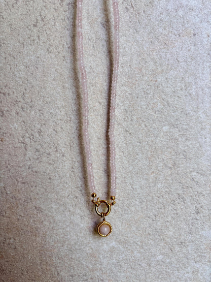 Rosenquarz-Perlenkette mit einem runden goldenen Rosenquarz Anhänger hängend vor grauer wand