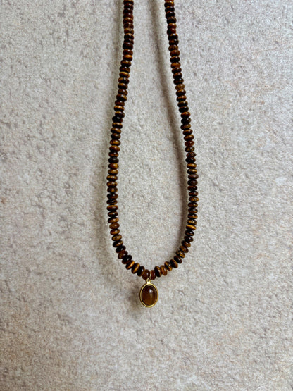 Braune Tigerauge-Perlenkette mit einem ovalen goldenen Tigerauge Anhänger hängend vor grauer Wand