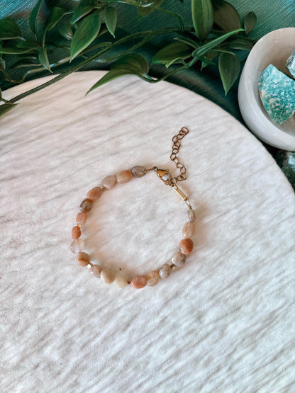 Sonnenstein-Armband Pearl auf einem weißen Stoff, umgeben von grünen Blättern und türkisfarbenen Steinen.