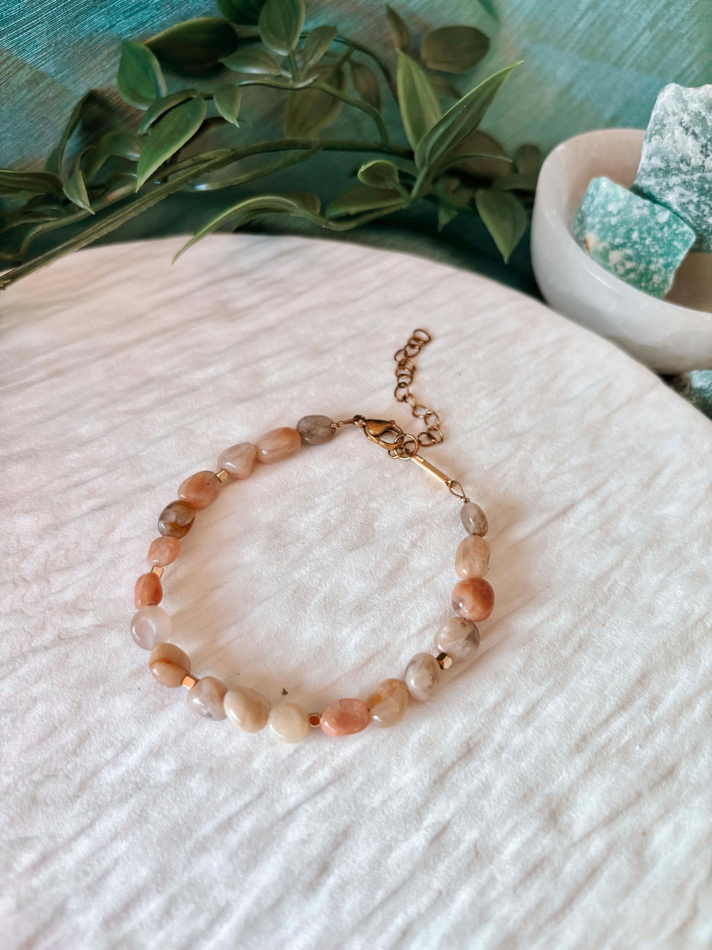 Sonnenstein-Armband "Pearl" auf hellem Untergrund, umgeben von grünen Zweigen und türkisfarbenen Steinen.