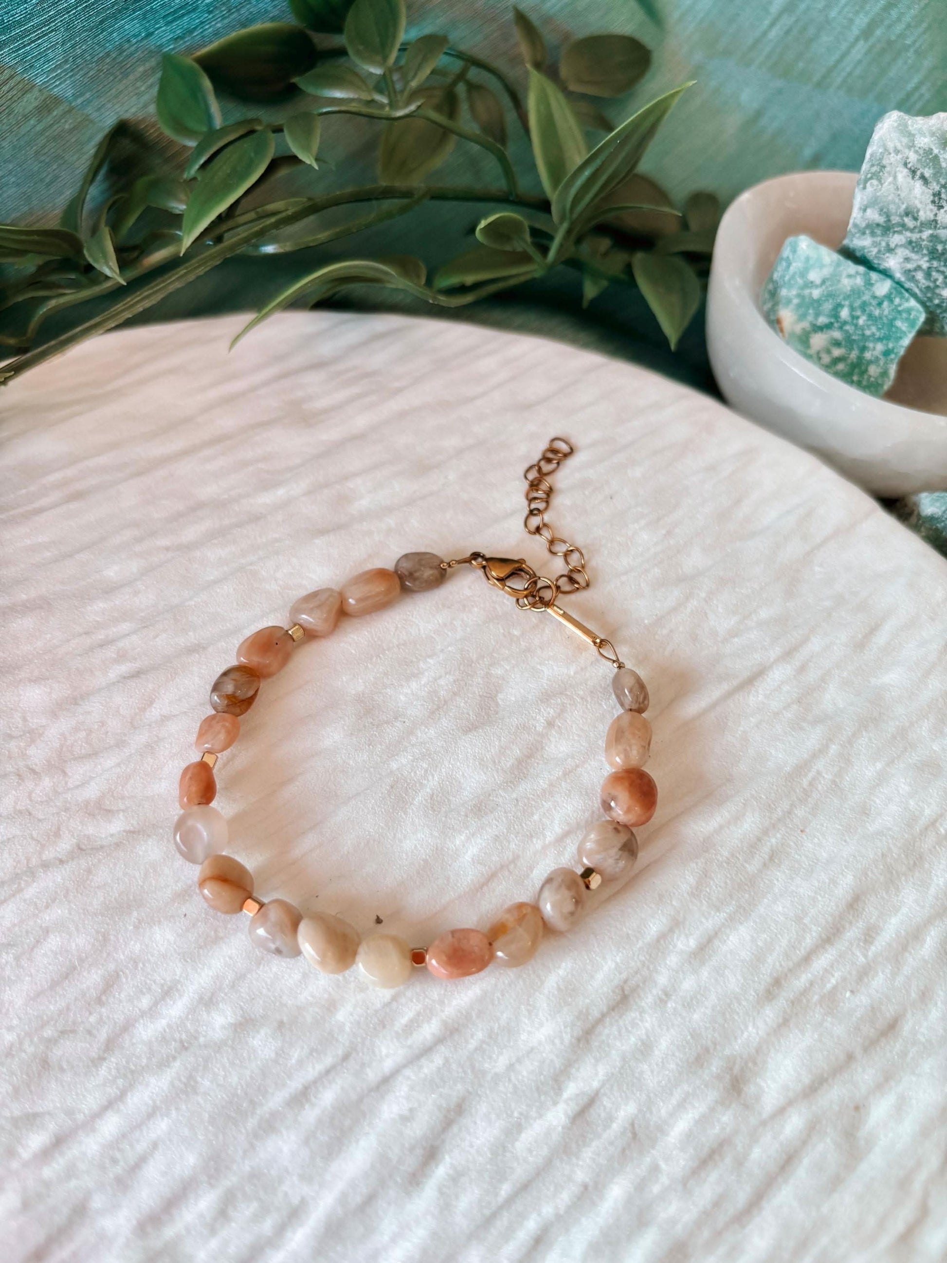 Sonnenstein-Armband "Pearl" auf hellem Untergrund, umgeben von grünen Zweigen und türkisfarbenen Steinen.