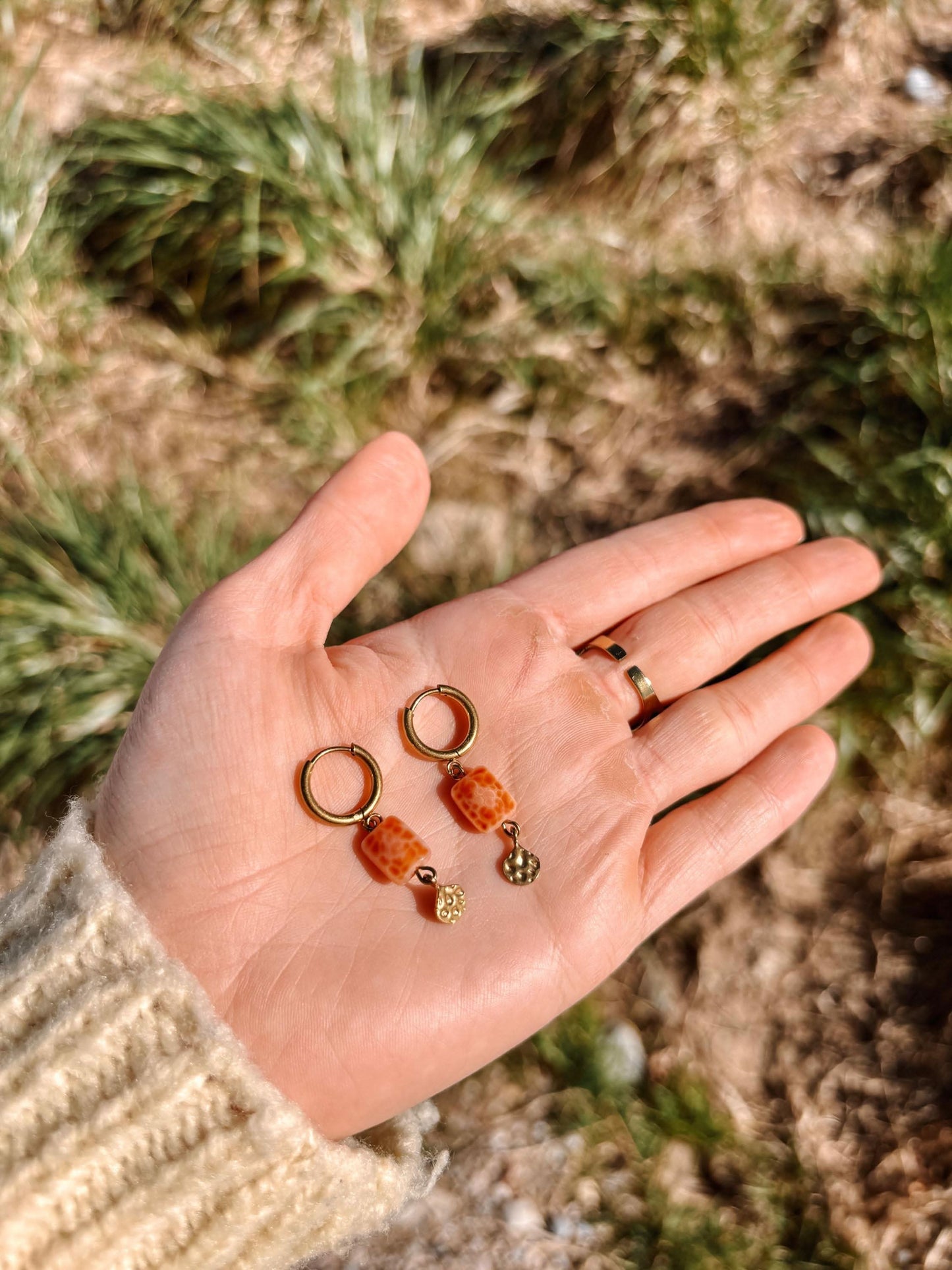Hand hält Achat-Ohrringe Moana mit orangenem Stein und Goldakzenten vor natürlichem Hintergrund.