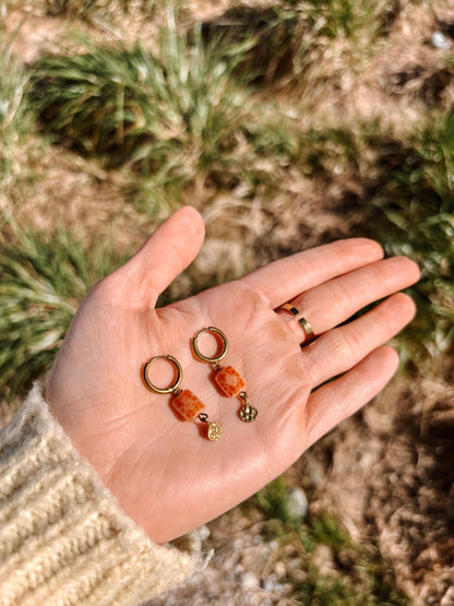 Hand hält Achat-Ohrringe Moana mit orangenem Stein und Goldakzenten vor natürlichem Hintergrund.
