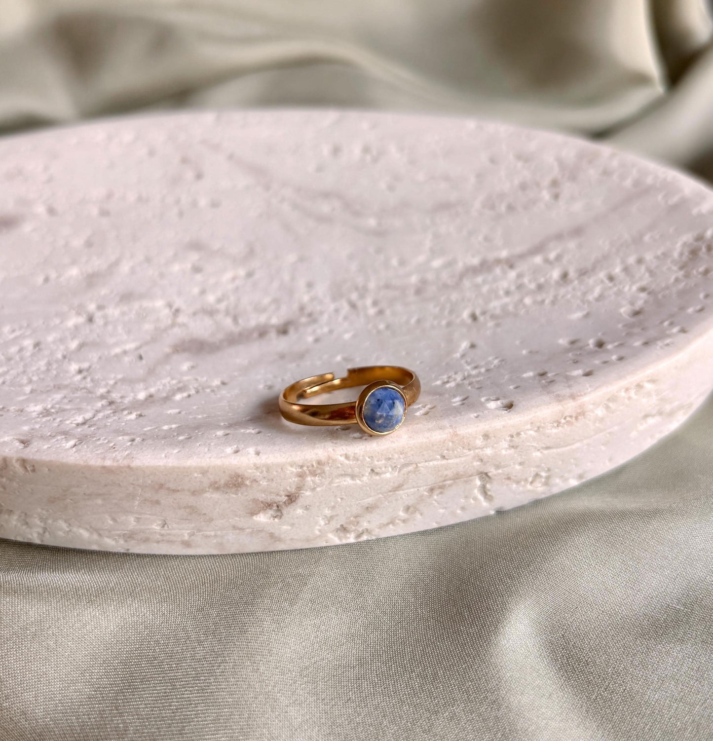 Goldener Ring mit einem runden Lapislazuli-Stein auf einer weißen, strukturierten Platte liegend.