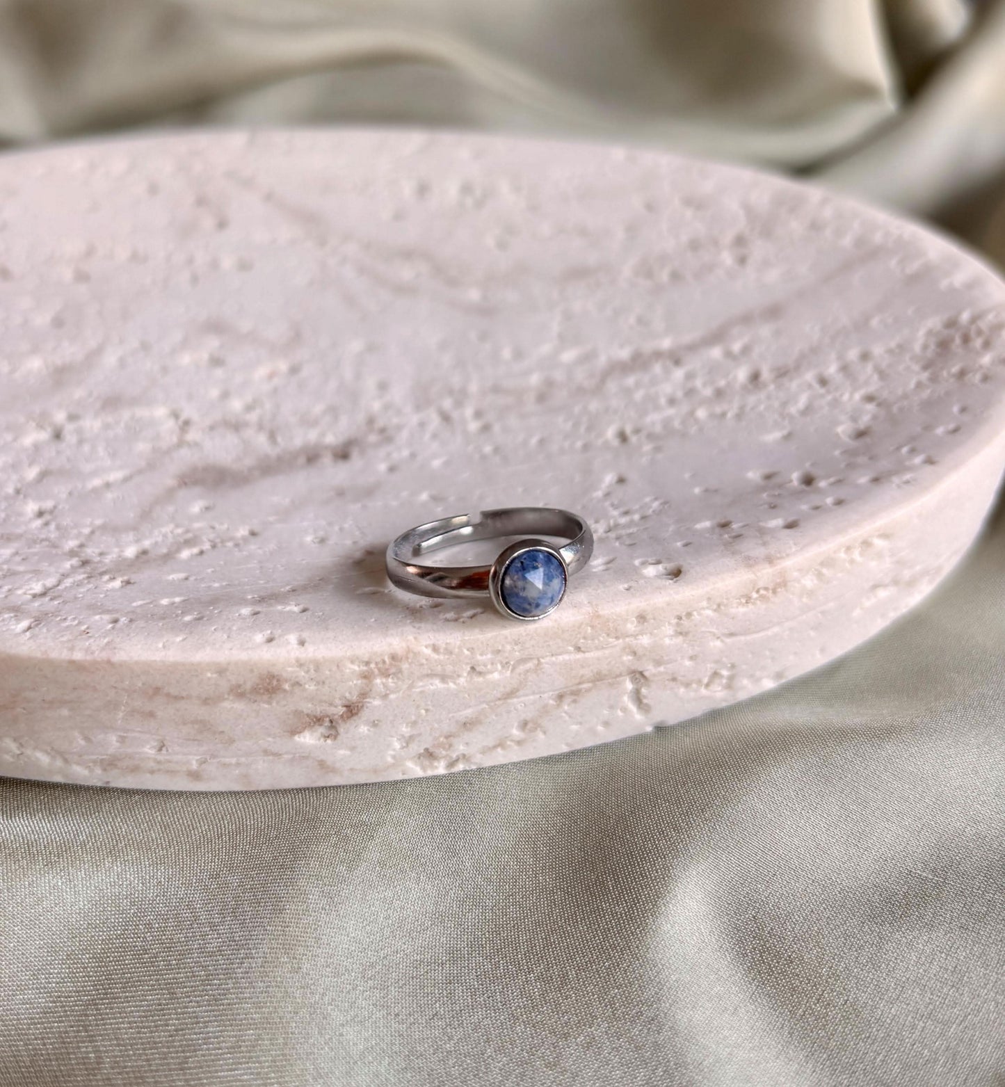 Silberner Ring mit einem Lapislazuli-Stein auf einem strukturierten weißen Teller, Hintergrund aus beigem Stoff.